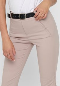 Vaalean beige vyötäröstä istuvat housut, jotka on valmistettu joustavasta kankaasta. Niissä on etutaskut ja musta nahkavyö, jossa on metallinen solki.