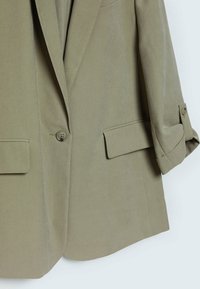 Blazer verde oliva realizzato in tessuto liscio con chiusura a un bottone, due tasche frontali e maniche arrotolate. Taglio sartoriale con linee pulite.
