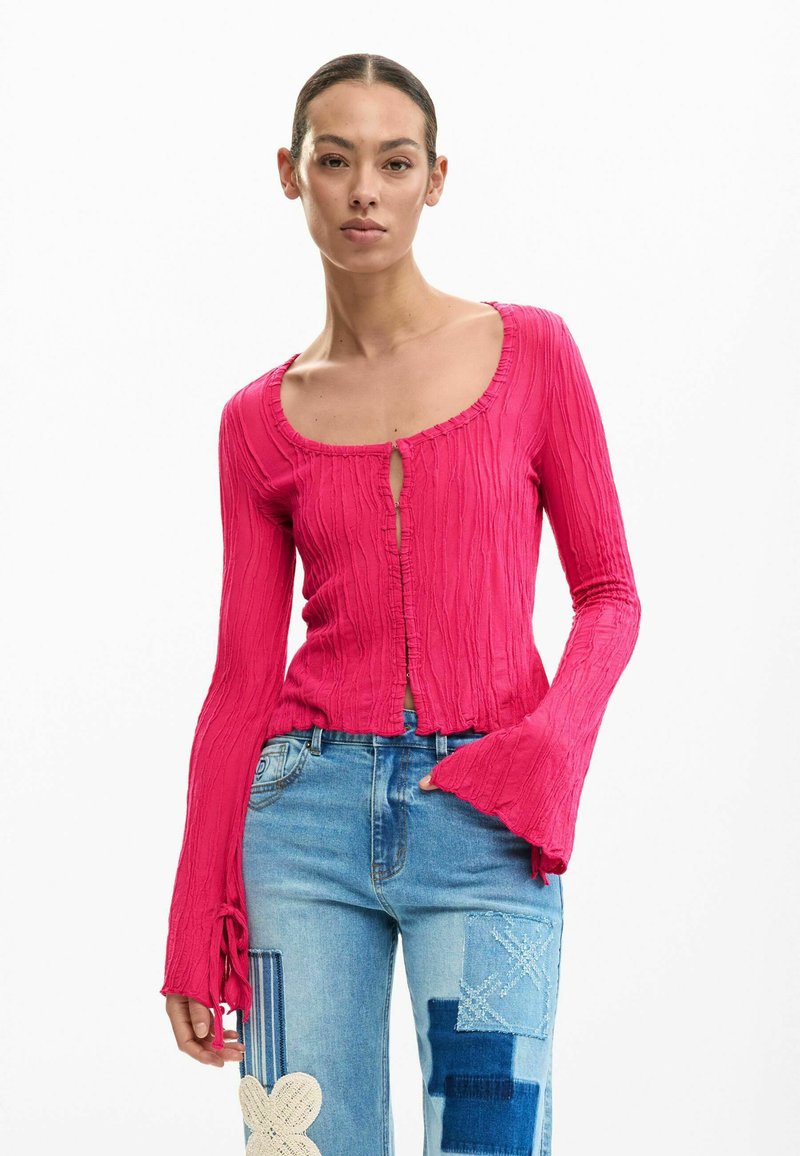 Femme portant un haut à manches longues texturé rose vif avec des poignets évasés et un jean patchwork bleu, debout devant un fond blanc.