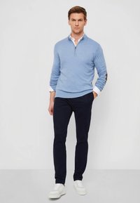 Pulôver azul claro com zíper no pescoço, mangas longas, barra canelada, combinado com calças azul-marinho e ténis brancos. Textura suave, design casual.