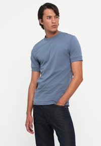 Men's blue short-sleeve t-shirt in soft cotton, featuring a crew neck and rolled cuffs, paired with dark denim jeans.
