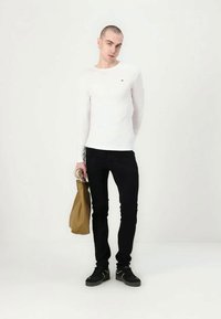 Mlada osoba s kratkom kosom koja nosi bijelu majicu dugih rukava, crne hlače, crne cipele i drži smotanu bež torbu uz jednostavnu pozadinu.