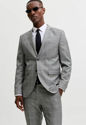 Homme portant un costume à carreaux gris avec une cravate noire et des lunettes de soleil, debout avec une main dans la poche devant un fond uni.