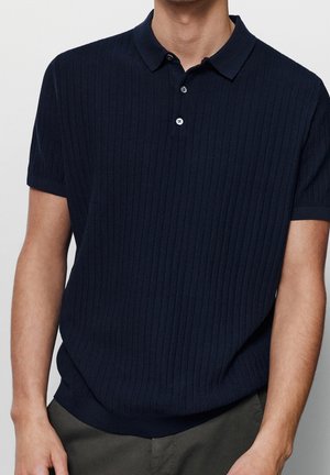 Mand iført en marineblå tekstureret kortærmet poloshirt med to-knaps slids, kombineret med mørkegrå bukser.