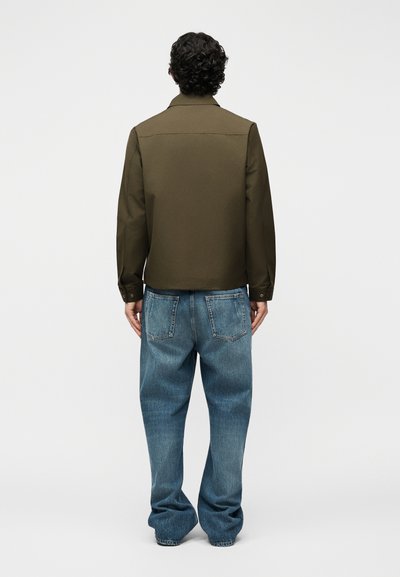 Homme de dos portant une veste vert olive et un jean bleu ample sur un fond clair uni.
