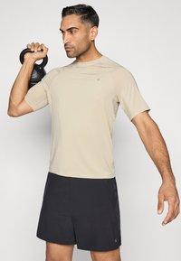 Világos bézs színű rövid ujjú sportpóló, fekete sorttal párosítva. A személy egy fekete kettlebellt tart. Simább anyagú felület.