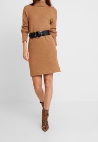 Robe en tricot beige avec un col montant, taille cintrée par une ceinture noire texturée. Assortie à des bottines à imprimé serpent marron.