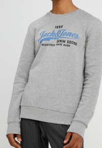 Hombre vestido con un sudadera de color gris claro que lleva el texto "1990 Jack & Jones Denim Goods Registered Trade Mark" en azul y negro en el pecho.