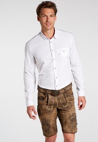 Camisa blanca de botones con mangas largas; pantalones cortos de cuero marrón con costuras decorativas y bolsillos; acento de hebilla tradicional en el cinturón.
