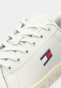 Hvite sneakers laget av glatt syntetisk materiale med runde snøringer. Har en logoetikett i svart, rødt og hvitt samt preget merkevare på sålen.