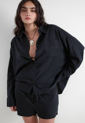 Junge Frau mit dunklen Haaren trägt ein übergroßes schwarzes Hemd mit Knöpfen, passende Shorts und eine Anhänger-Halskette, posiert vor einem weißen Hintergrund.