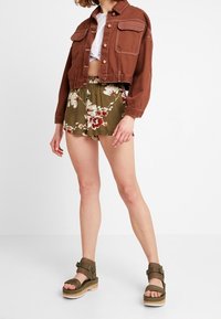 Veste en denim marron cropped avec poches à rabat, crop top blanc, shorts fleuris vert olive et sandales à plateforme vert olive.