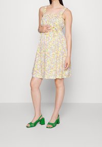 Kwiatowa sukienka na żółtym tle, kwadratowy dekolt i falbaniaste ramiączka. Zestawiona z zielonymi sandałami na obcasie z wycięciami.