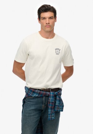 Camiseta de algodón color crema con un logo azul de "SUPER DRY" en el pecho. Combinada con jeans azules y una camisa de cuadros atada a la cintura.