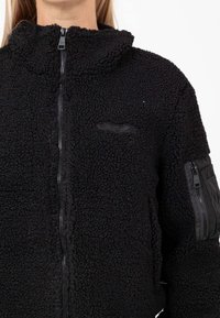 Giacca in pile nera con colletto alto, chiusura con zip, superficie texturizzata e un piccolo logo sul petto. Presenta una tasca con zip sulla manica.