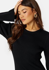 Maglione nero a coste con maniche a campana lunghe; scollatura rotonda. Il tessuto ha un aspetto texturizzato, che mette in risalto il motivo a maglia. Orecchini a cerchio dorati.