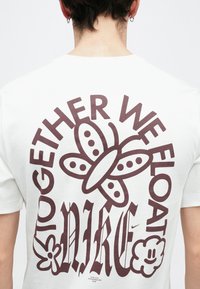 Biały bawełniany t-shirt z dużym brązowym nadrukiem z tekstem "RAZEM UNOSIMY SIĘ" oraz motylem i kwiatowymi akcentami.