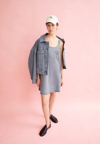 Jeansjack met grote zakken over een grijze denim jurk. Zwarte instappers. Lichte achtergrond met zachte roze tonen.