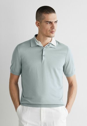 Mann mit kurzen Haaren, der ein hellgrünes Polohemd und weiße Hose trägt, steht mit den Händen in den Taschen und schaut nach rechts vor einem einfarbigen Hintergrund.