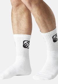 Jambes portant des chaussettes blanches côtelées avec un logo abstrait noir sur les côtés, sur un fond clair et uni.