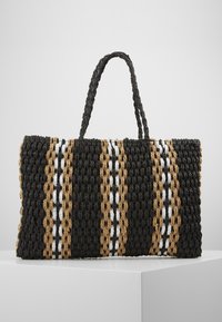 Woven tote bag o prostokątnym kształcie, z czarnymi, brązowymi i białymi paskami, teksturowaną powierzchnią oraz dwiema plecionymi rączkami.