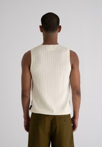 Cream ribbed knit sleeveless top with a round neck, paired with dark olive green trousers featuring button accents on the back pockets.