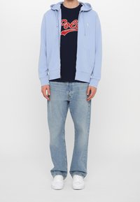 Ljusblå zip-hoodie med ficka fram, marinblå t-shirt med grafiskt tryck som har röd text, ljusblå jeans och vita sneakers.