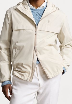 Mann trägt eine beige Reißverschlussjacke über einem blauen Strickpullover und einem weißen Hemd mit Kragen, weiße Hose, Hände in den Taschen.