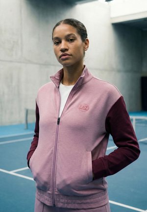 Donna che indossa una giacca sportiva con zip rosa e bordeaux, in piedi su un campo da tennis coperto blu con le mani in tasca.