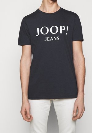 Mand iført en sort t-shirt med korte ærmer med hvid tekst "JOOP! JEANS" og hvide bukser, stående mod en ensfarvet baggrund.