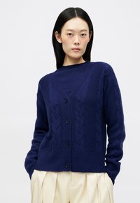 Σκούρο μπλε κάρντινγκ με σχέδιο καμπύλης πλέξης, V λαιμόκοψη και κουμπιά μπροστά. Ριμπύ μανσέτες και στρίφωμα, κατασκευασμένο από μαλακό υλικό με υφή.