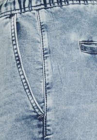 Vaaleansininen farkkukangas tekstuurilla, jossa on valkoista ommelta, sivutaskuja ja rento vyötärö.