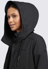 Veste à capuche noire avec une finition brillante, dotée de cordons de serrage ajustables et d'accents de logo. Le tissu semble lisse et léger.