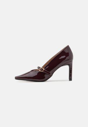 Chaussure à talon aiguille bordeaux en patent avec un embout pointu, une bride de cheville comportant des accents en métal doré, et une finition lisse et brillante. Talon stiletto surélevé.