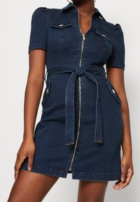 Vestido de mezclilla en azul oscuro, con cierre frontal, mangas cortas y dos bolsillos en el pecho. Incluye un cinturón atado en la cintura y sutiles detalles de costura.
