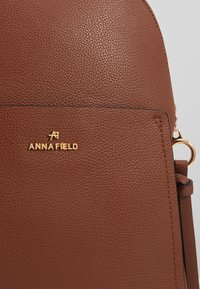 Zaino in pelle marrone testurizzata con logo dorato "ANNA FIELD." Presenta bordi lisci e una silhouette curva, con una tasca laterale e finiture dorate.