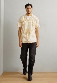 Krátky rukáv, béžová košeľa so zlatým kvetinovým vzorom, skombinovaná s tmavými džínsami a čiernymi čižmami. Model stojí na neutrálnom pozadí.