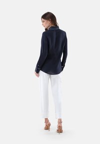 Marineblaue Leinenbluse mit Kragen, langen Ärmeln und weißen Details, kombiniert mit weißen, hochgekrempelten Hosen und beigen High-Heels.