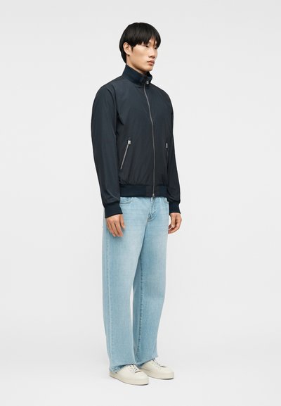 Άνδρας που φοράει μαύρο μπουφάν με φερμουάρ και τσέπες με φερμουάρ, ανοιχτόχρωμο τζιν παντελόνι με φαρδιές μπατζάκια και λευκά αθλητικά παπούτσια, στέκεται μπροστά σε απλό φόντο.