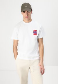 White cotton t-shirt with a multicolored graphic on the left chest. Paired with light beige trousers and an olive green baseball cap.