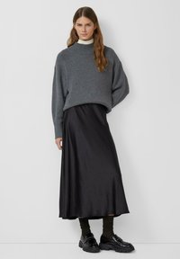 Grijze oversized trui, zwarte satijnen midi rok en glanzende zwarte schoenen. Getextureerde brei, relaxte pasvorm en gelaagde styling.