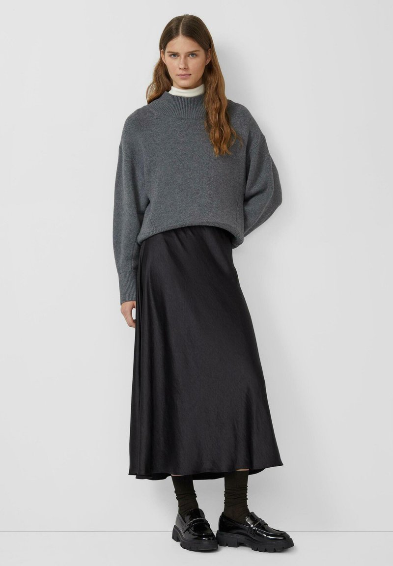 Grijze oversized trui, zwarte satijnen midi rok en glanzende zwarte schoenen. Getextureerde brei, relaxte pasvorm en gelaagde styling.