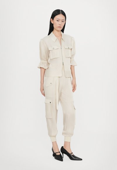 Beige kurze Jacke mit puffigen Ärmeln und vorderen Taschen, kombiniert mit passenden schmal geschnittenen Cargo-Hosen. Schwarze spitze Absätze runden das Outfit ab.