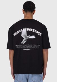 Svart bomulls t-skjorte med et hvitt duegrafikk og teksten "PEACE IN PARADISE" på baksiden, med ekstra motiverende tekst under.