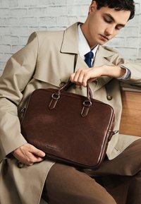 Brun læder laptop taske med dobbelt håndtag og sølvhardware. Glat tekstur, rektangulær form. Minimalistisk design med prægning af brand.
