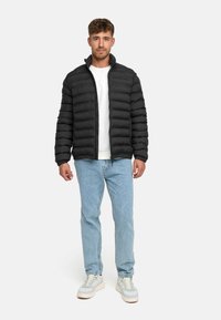 Sort pufferjakke med vandret kviltet design, høj krave og frontlås. Kombineret med lyseblå jeans og afslappede sneakers.