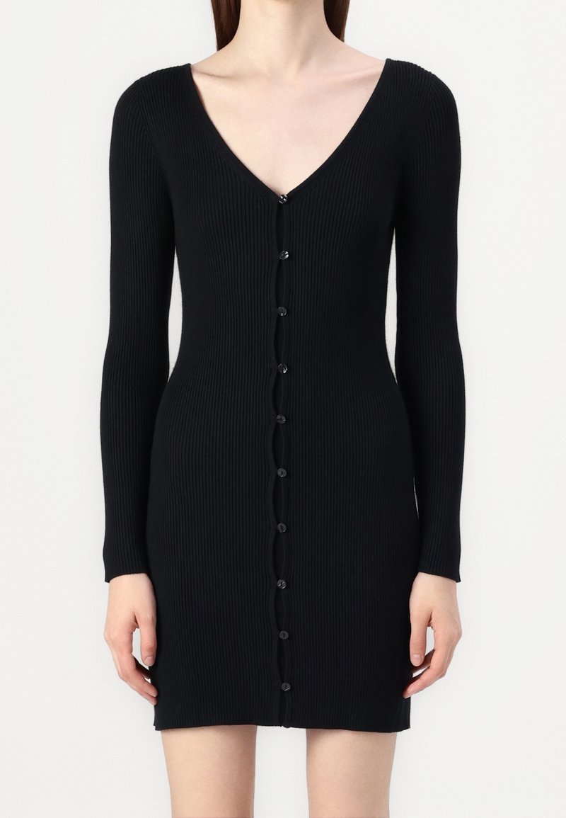Femme portant une robe en maille côtelée noire ajustée avec manches longues, col en V profond et fermeture à boutons sur le devant, du col à l'ourlet.