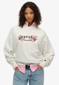 Felpa beige chiaro con ricami floreali e testo nero "Super Los Angeles", sovrapposta a una maglietta a righe rosa. Vestibilità casual, polsini a coste.
