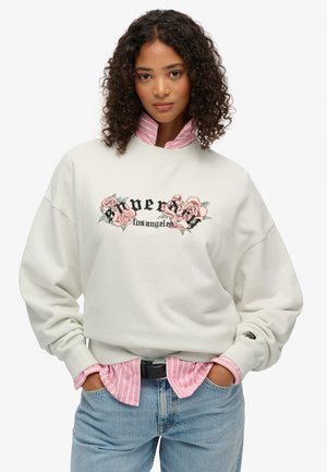 Sweatshirt beige clair avec broderies florales et texte noir "Super Los Angeles", superposé à une chemise à rayures rose. Coupe décontractée, poignets côtelés.