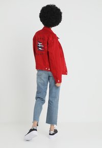 Veste en denim rouge avec un détail de logo au dos, assortie à un jean droit bleu clair et des baskets noir et blanc.